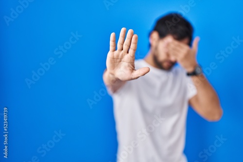 Handsome latin man standing over blue background covering eyes with hands and doing stop gesture with sad and fear expression. embarrassed and negative concept.