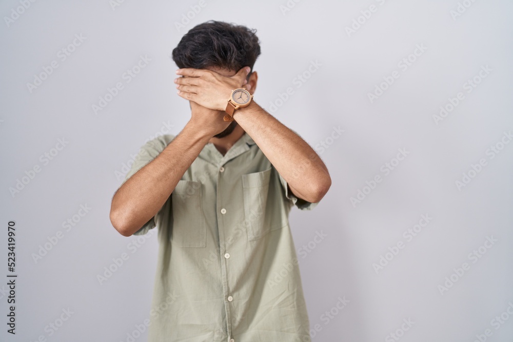 Arab man with beard standing over white background covering eyes and mouth with hands, surprised and shocked. hiding emotion Arab man with beard standing over white background covering eyes and mouth with hands, surprised and shocked. hiding emotion