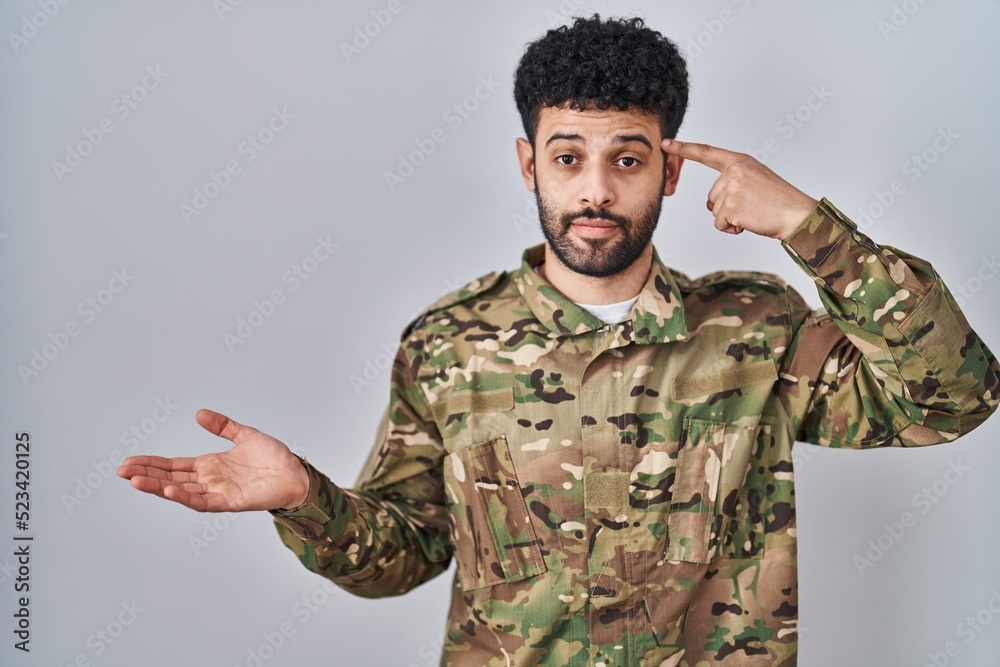 Arab man wearing camouflage army uniform confused and annoyed with open palm showing copy space and pointing finger to forehead. think about it. Arab man wearing camouflage army uniform confused and annoyed with open palm showing copy space and pointing finger to forehead. think about it.