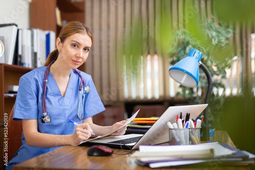 Young woman professional physician filling up medical forms on laptop while sitting at table in office..