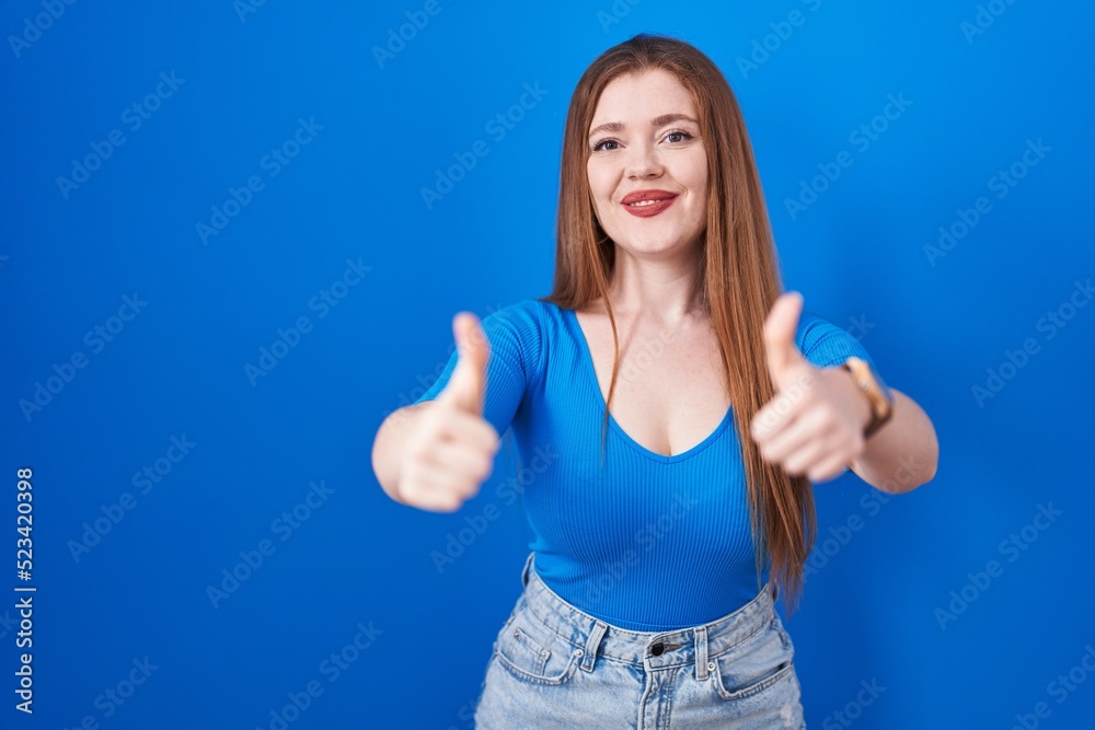 Redhead woman standing over blue background approving doing positive gesture with hand, thumbs up smiling and happy for success. winner gesture.