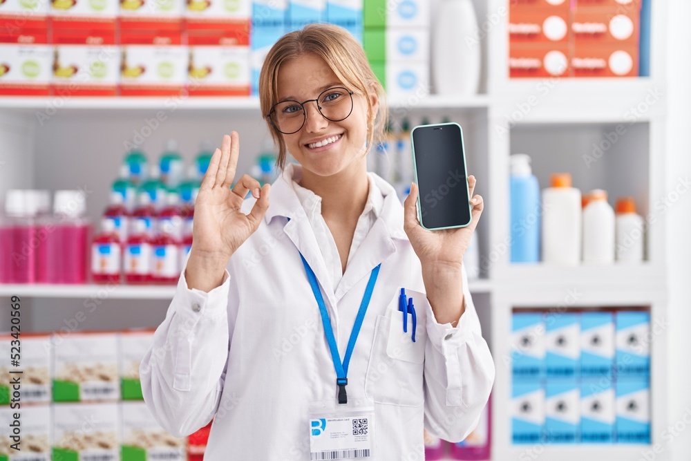 Young caucasian woman working at pharmacy drugstore showing smartphone screen doing ok sign with fingers, smiling friendly gesturing excellent symbol