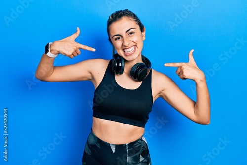 Beautiful hispanic woman wearing gym clothes and using headphones looking confident with smile on face, pointing oneself with fingers proud and happy.