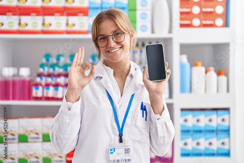 Young caucasian woman working at pharmacy drugstore showing smartphone screen doing ok sign with fingers  smiling friendly gesturing excellent symbol