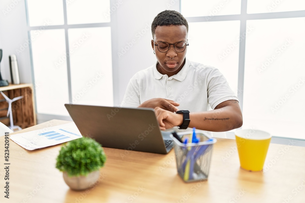 Young african man working at the office using computer laptop checking the time on wrist watch, relaxed and confident