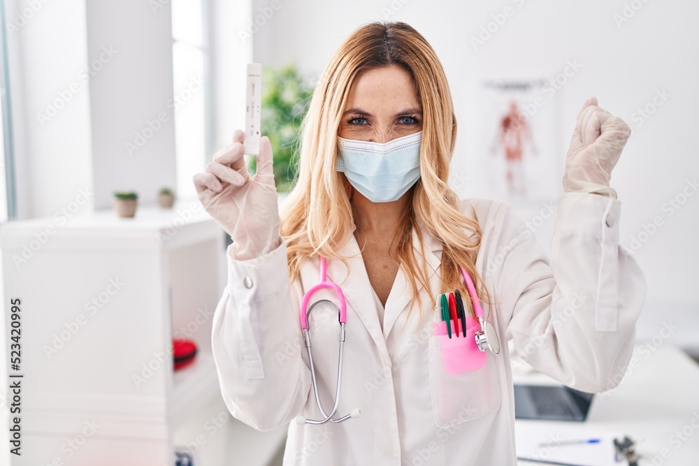 Young caucasian doctor woman holding covid test screaming proud, celebrating victory and success very excited with raised arms Young caucasian doctor woman holding covid test screaming proud, celebrating victory and success very excited with raised arms