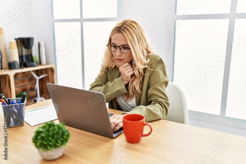 Beautiful blonde woman working at the office with laptop looking confident at the camera with smile with crossed arms and hand raised on chin. thinking positive.