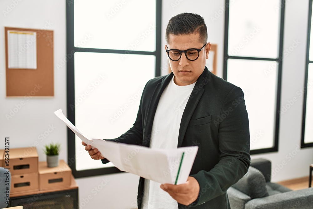 Young latin businessman with serious expression reading paperwork standing at the office.
