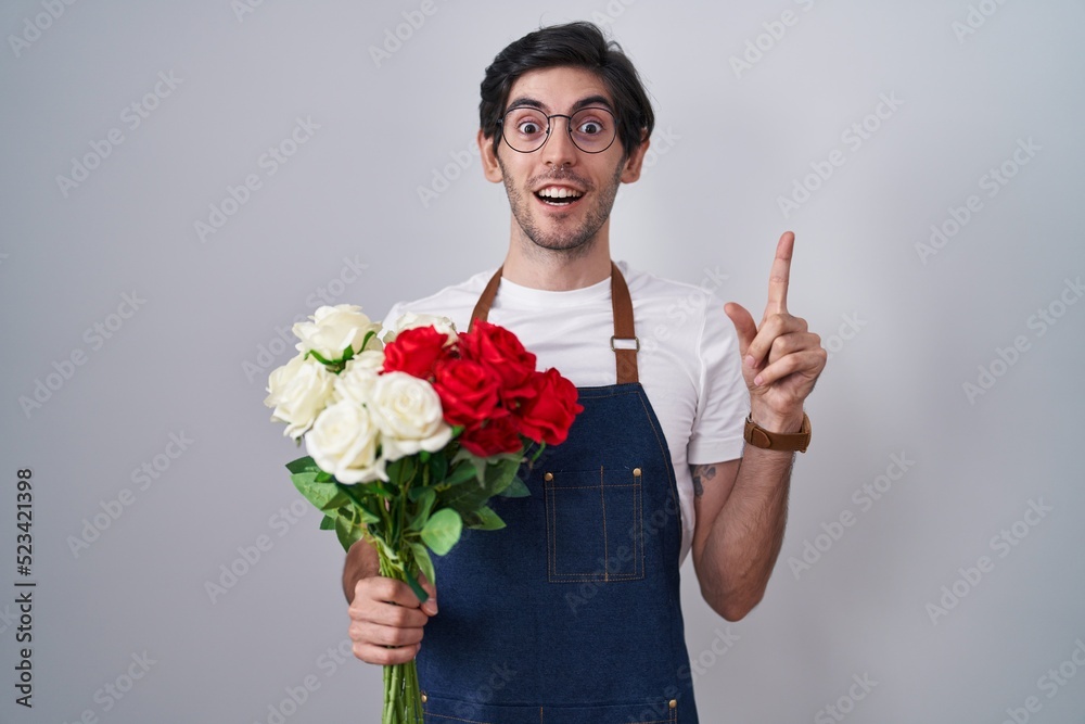 Young hispanic man holding bouquet of white and red roses pointing finger up with successful idea. exited and happy. number one. Young hispanic man holding bouquet of white and red roses pointing finger up with successful idea. exited and happy. number one.