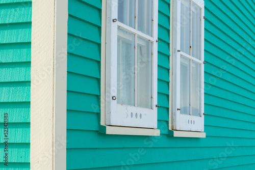 Two double hung windows on a green exterior wall of a vintage style building. The building has narrow horizontal clapboard siding. There s white wooden shutter and trim around the closed glass window