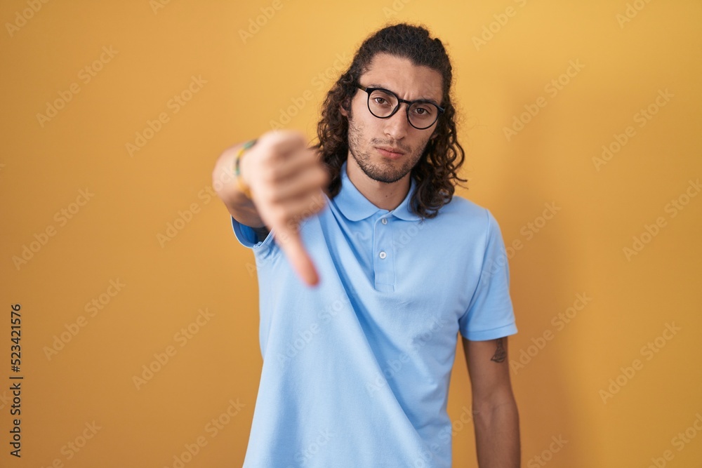 Young hispanic man standing over yellow background looking unhappy and angry showing rejection and negative with thumbs down gesture. bad expression. Young hispanic man standing over yellow background looking unhappy and angry showing rejection and negative with thumbs down gesture. bad expression.
