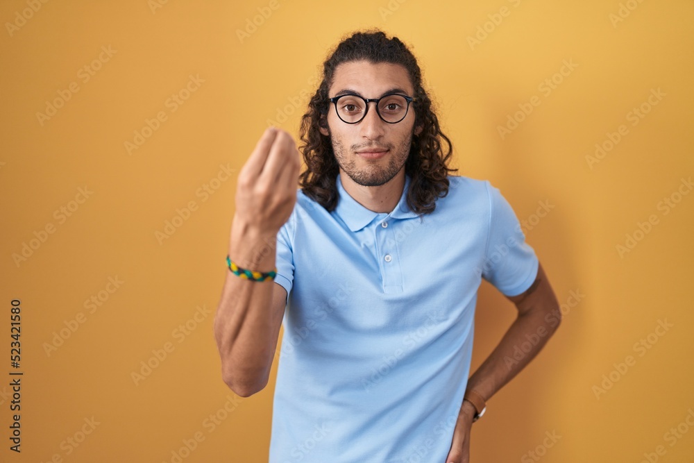 Young hispanic man standing over yellow background doing italian gesture with hand and fingers confident expression Young hispanic man standing over yellow background doing italian gesture with hand and fingers confident expression