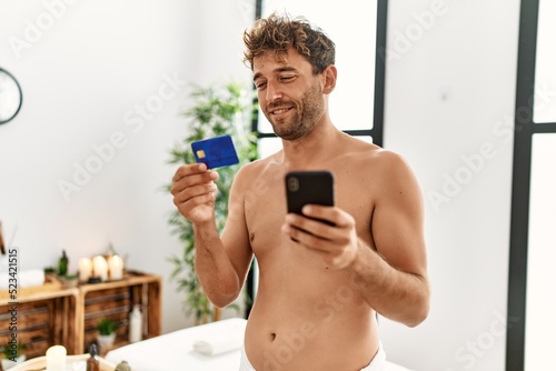 Young hispanic man smiling confident holding credit card and smartphone at beauty center