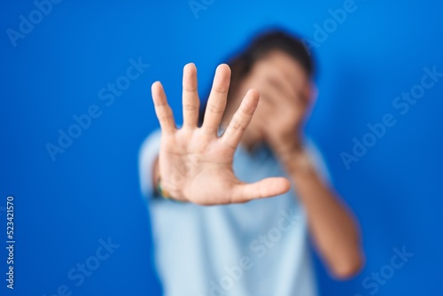 Young hispanic man standing over blue background covering eyes with hands and doing stop gesture with sad and fear expression. embarrassed and negative concept.