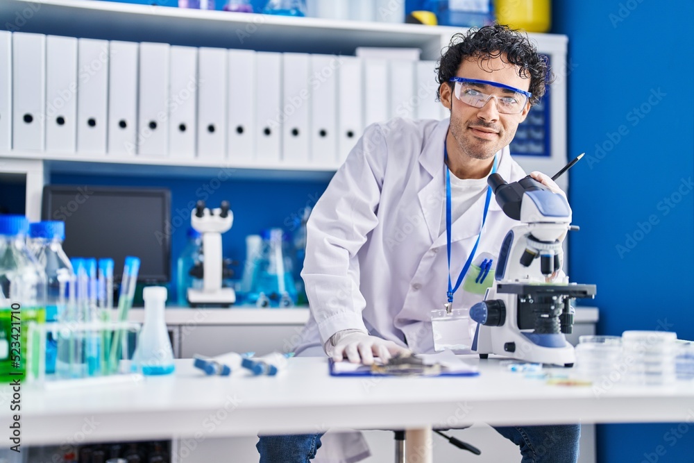 Fototapeta premium Young hispanic man scientist writing on document using microscope at laboratory