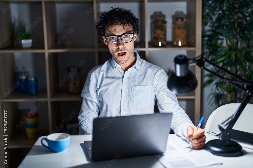 Obraz premium Hispanic man working at the office at night in shock face, looking skeptical and sarcastic, surprised with open mouth