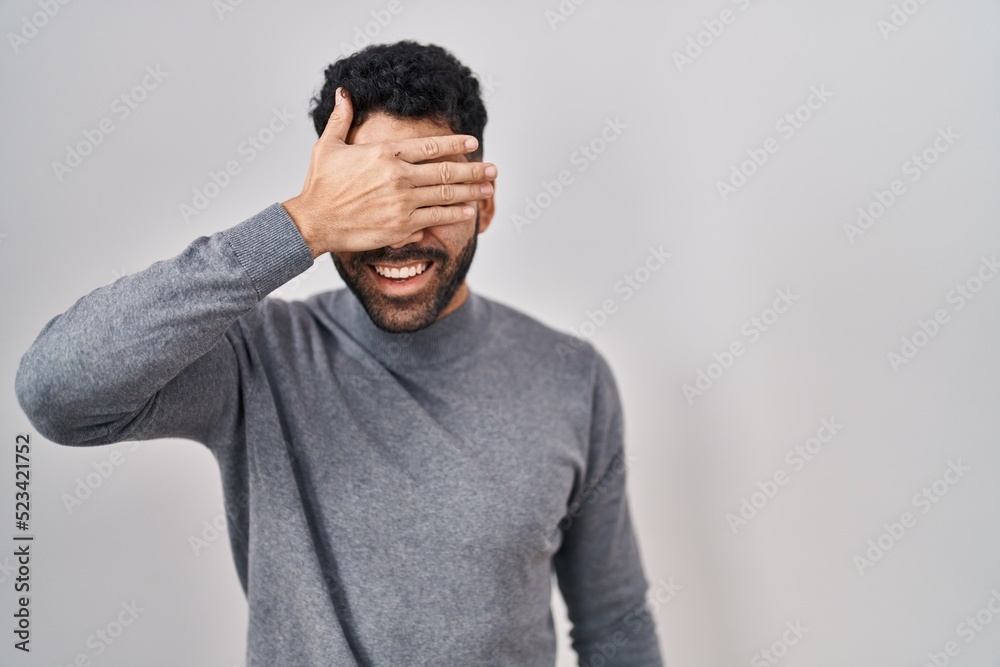 Hispanic man with beard standing over white background smiling and laughing with hand on face covering eyes for surprise. blind concept. Hispanic man with beard standing over white background smiling and laughing with hand on face covering eyes for surprise. blind concept.