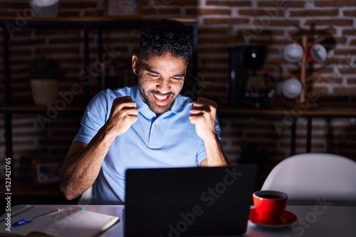 Hispanic man with beard using laptop at night very happy and excited doing winner gesture with arms raised, smiling and screaming for success. celebration concept.