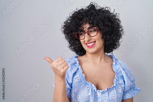 Young brunette woman with curly hair wearing glasses over isolated background smiling with happy face looking and pointing to the side with thumb up.