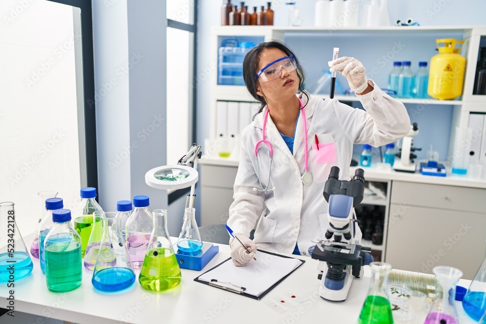Young chinese woman wearing scientist uniform holding blood test tube writing on document at laboratory Young chinese woman wearing scientist uniform holding blood test tube writing on document at laboratory