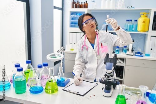 Young chinese woman wearing scientist uniform holding blood test tube writing on document at laboratory