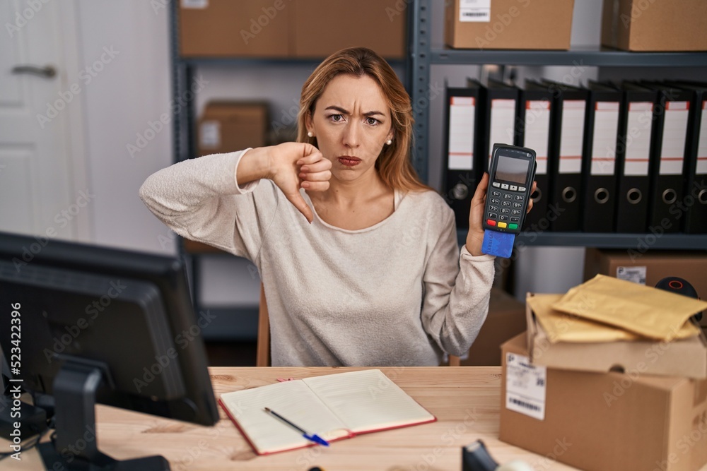 Hispanic woman working at small business ecommerce holding credit card and dataphone with angry face, negative sign showing dislike with thumbs down, rejection concept Hispanic woman working at small business ecommerce holding credit card and dataphone with angry face, negative sign showing dislike with thumbs down, rejection concept