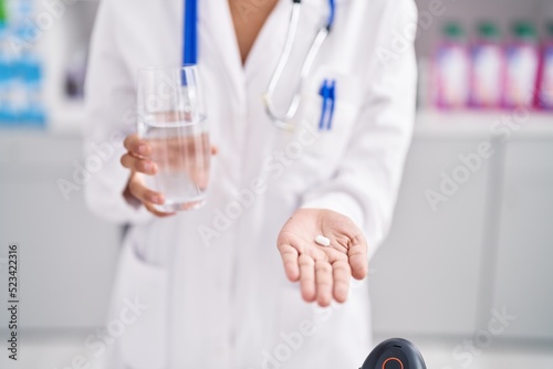 Young hispanic woman pharmacist holding pills and glass of water at pharmacy