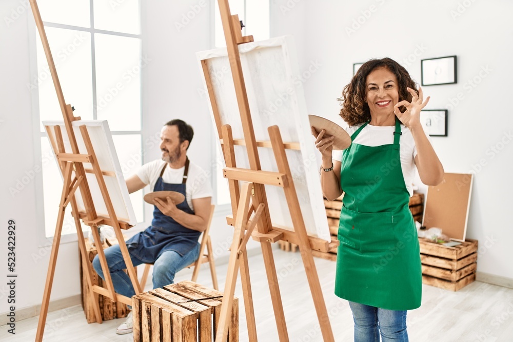 Hispanic middle age woman and mature man at art studio doing ok sign with fingers, smiling friendly gesturing excellent symbol