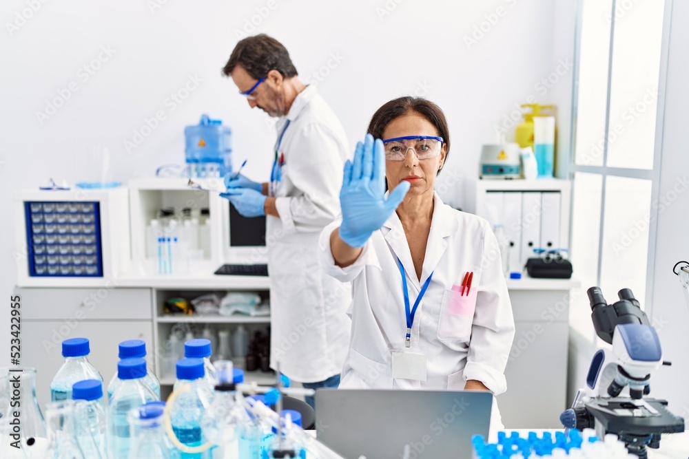 Middle age woman working at scientist laboratory with open hand doing stop sign with serious and confident expression, defense gesture Middle age woman working at scientist laboratory with open hand doing stop sign with serious and confident expression, defense gesture