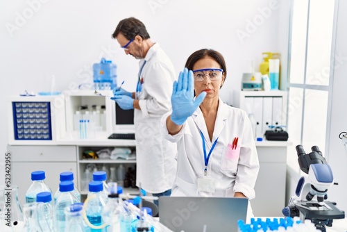 Middle age woman working at scientist laboratory with open hand doing stop sign with serious and confident expression, defense gesture