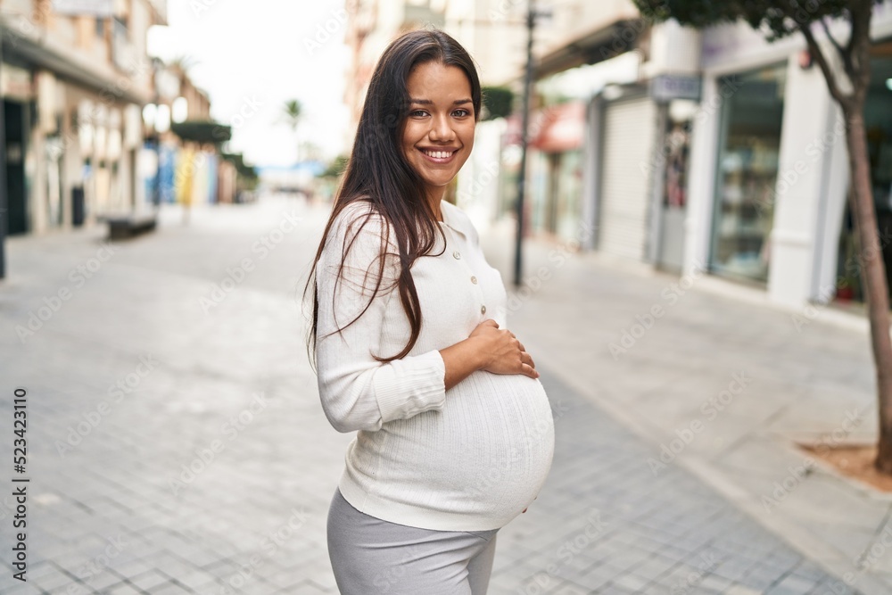 Young latin woman pregnant smiling confident touching belly at street