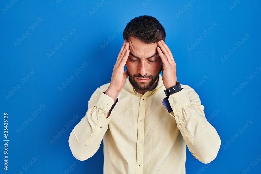 Handsome hispanic man standing over blue background with hand on head, headache because stress. suffering migraine. Handsome hispanic man standing over blue background with hand on head, headache because stress. suffering migraine.