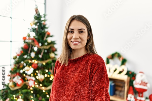 Young hispanic girl smiling happy standing by christmas tree at home.