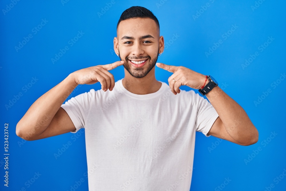 Young hispanic man standing over blue background smiling cheerful showing and pointing with fingers teeth and mouth. dental health concept.