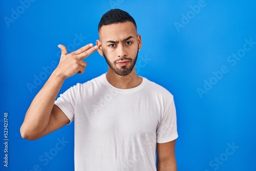 Young hispanic man standing over blue background shooting and killing oneself pointing hand and fingers to head like gun, suicide gesture. © Krakenimages.com