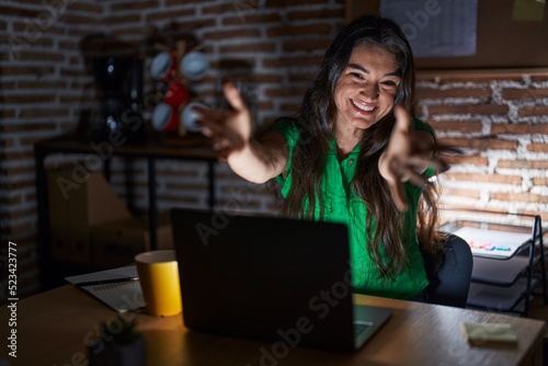 Young teenager girl working at the office at night looking at the camera smiling with open arms for hug. cheerful expression embracing happiness.