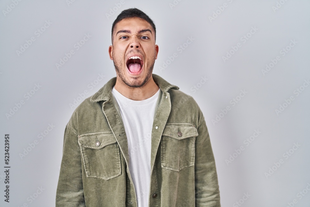 Young hispanic man standing over isolated background angry and mad screaming frustrated and furious, shouting with anger. rage and aggressive concept. Young hispanic man standing over isolated background angry and mad screaming frustrated and furious, shouting with anger. rage and aggressive concept.