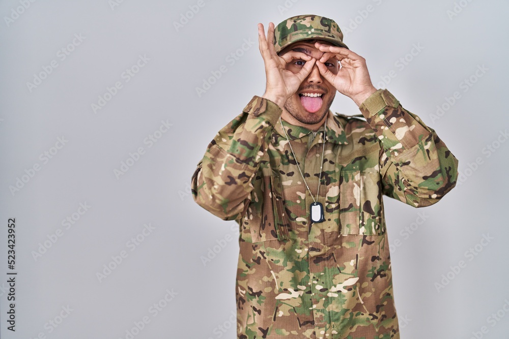 Young hispanic man wearing camouflage army uniform doing ok gesture like binoculars sticking tongue out, eyes looking through fingers. crazy expression. Young hispanic man wearing camouflage army uniform doing ok gesture like binoculars sticking tongue out, eyes looking through fingers. crazy expression.