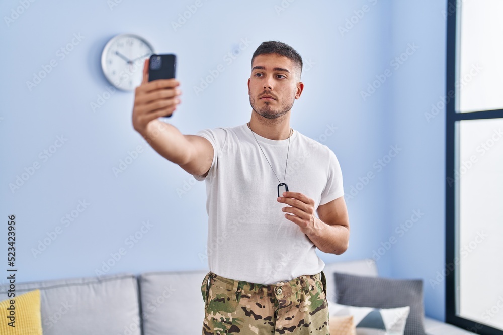 Young hispanic man wearing camouflage army uniform taking selfie at home relaxed with serious expression on face. simple and natural looking at the camera. Young hispanic man wearing camouflage army uniform taking selfie at home relaxed with serious expression on face. simple and natural looking at the camera.