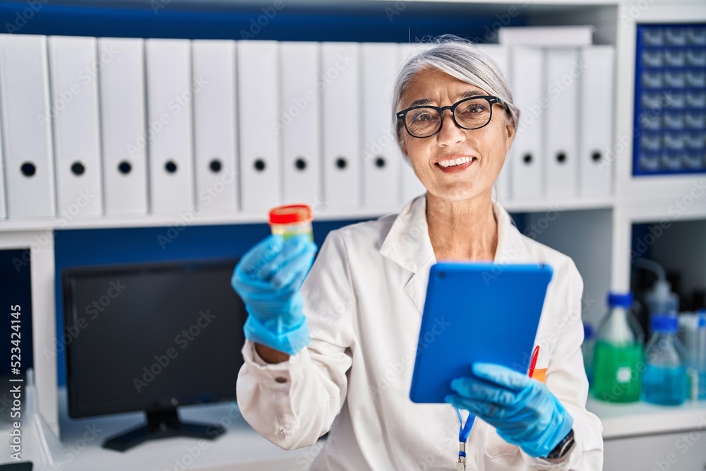 Middle age grey-haired woman scientist using touchpad holding test tube at laboratory Middle age grey-haired woman scientist using touchpad holding test tube at laboratory
