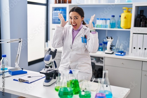 Young brunette woman working at scientist laboratory celebrating victory with happy smile and winner expression with raised hands