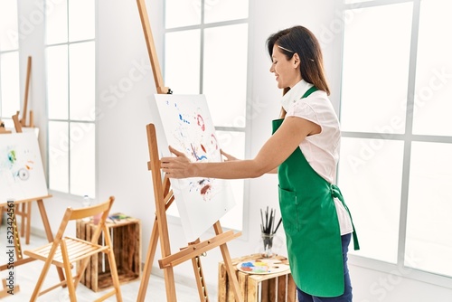 Middle age hispanic woman smiling confident holding canvas draw at art studio