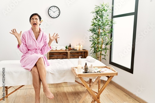 Beautiful caucasian woman wearing bathrobe at wellness spa crazy and mad shouting and yelling with aggressive expression and arms raised. frustration concept.