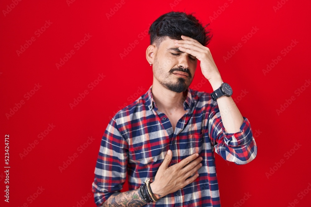 Young hispanic man with beard standing over red background touching forehead for illness and fever, flu and cold, virus sick Young hispanic man with beard standing over red background touching forehead for illness and fever, flu and cold, virus sick