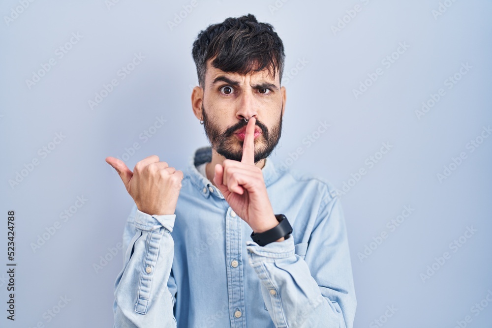Young hispanic man with beard standing over blue background asking to be quiet with finger on lips pointing with hand to the side. silence and secret concept. Young hispanic man with beard standing over blue background asking to be quiet with finger on lips pointing with hand to the side. silence and secret concept.