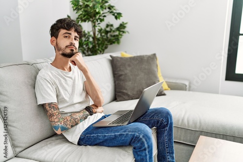 Hispanic man with beard sitting on the sofa with hand on chin thinking about question, pensive expression. smiling with thoughtful face. doubt concept.
