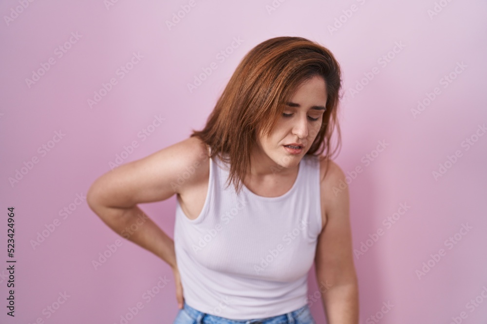 Brunette woman standing over pink background suffering of backache, touching back with hand, muscular pain Brunette woman standing over pink background suffering of backache, touching back with hand, muscular pain