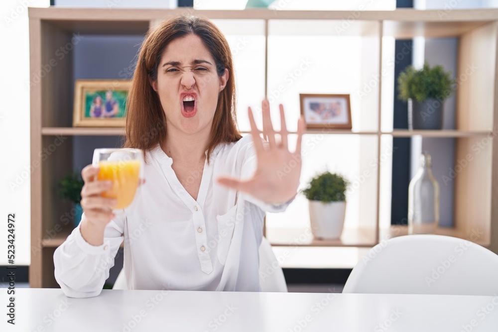 Brunette woman drinking glass of orange juice doing stop gesture with hands palms, angry and frustration expression Brunette woman drinking glass of orange juice doing stop gesture with hands palms, angry and frustration expression