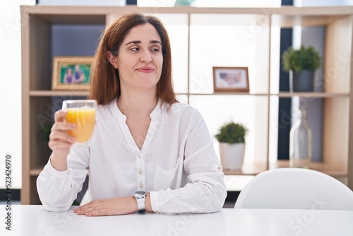 Brunette woman drinking glass of orange juice looking away to side with smile on face, natural expression. laughing confident.