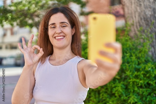 Brunette woman taking a selfie photo with smartphone doing ok sign with fingers, smiling friendly gesturing excellent symbol